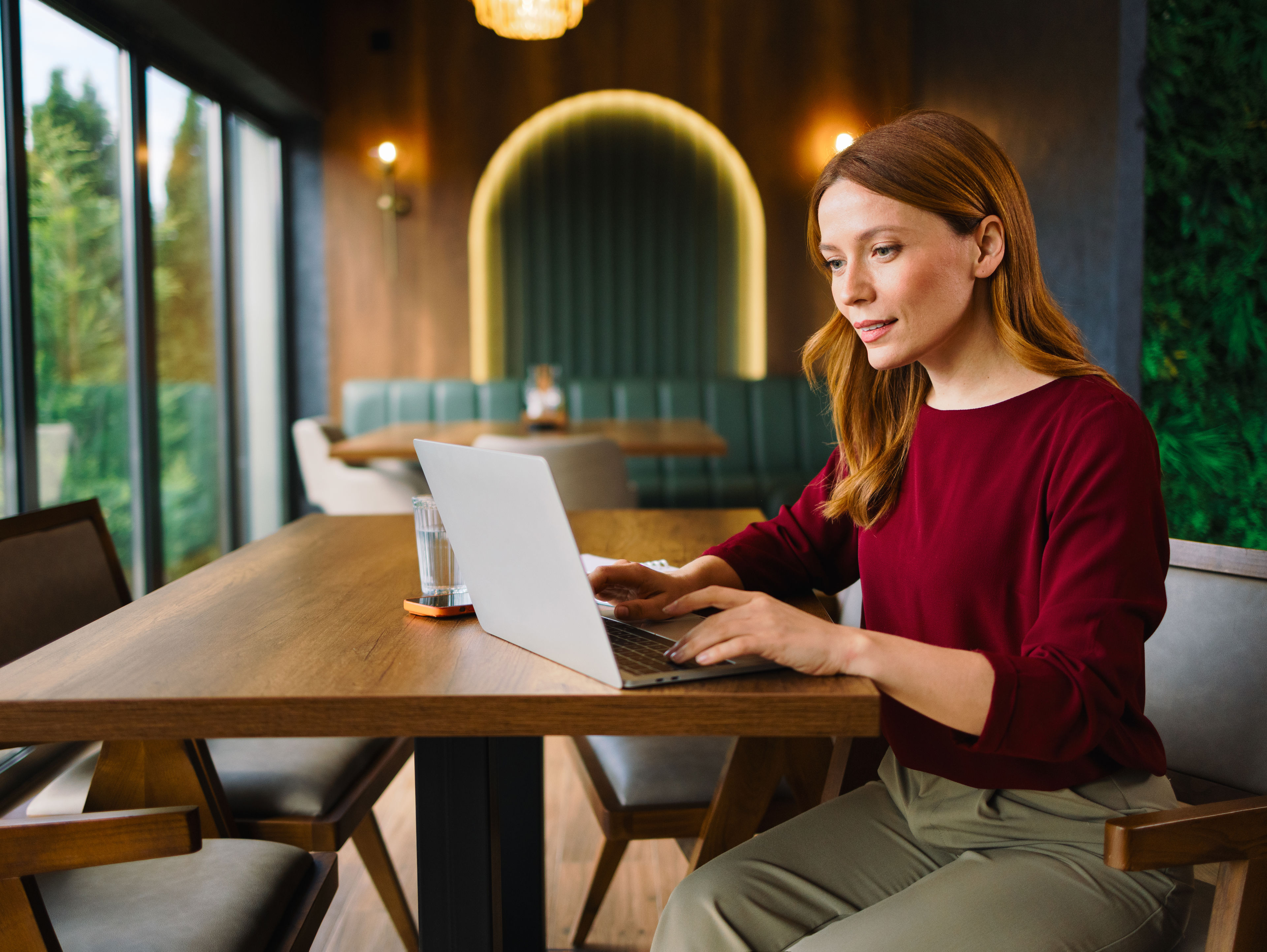 Woman with long red hair works on a laptop at a wooden table in a modern, well-lit cafe with large windows, green wall, and cozy booth seating in the background.