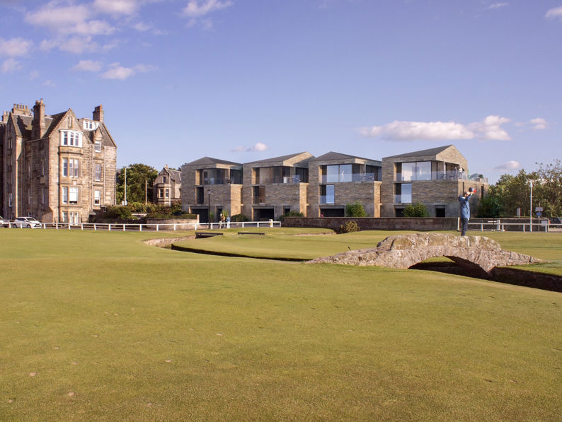A stone bridge crosses a golf course with modern buildings on the right and a historic stone building on the left, under a blue sky scattered with a few clouds.