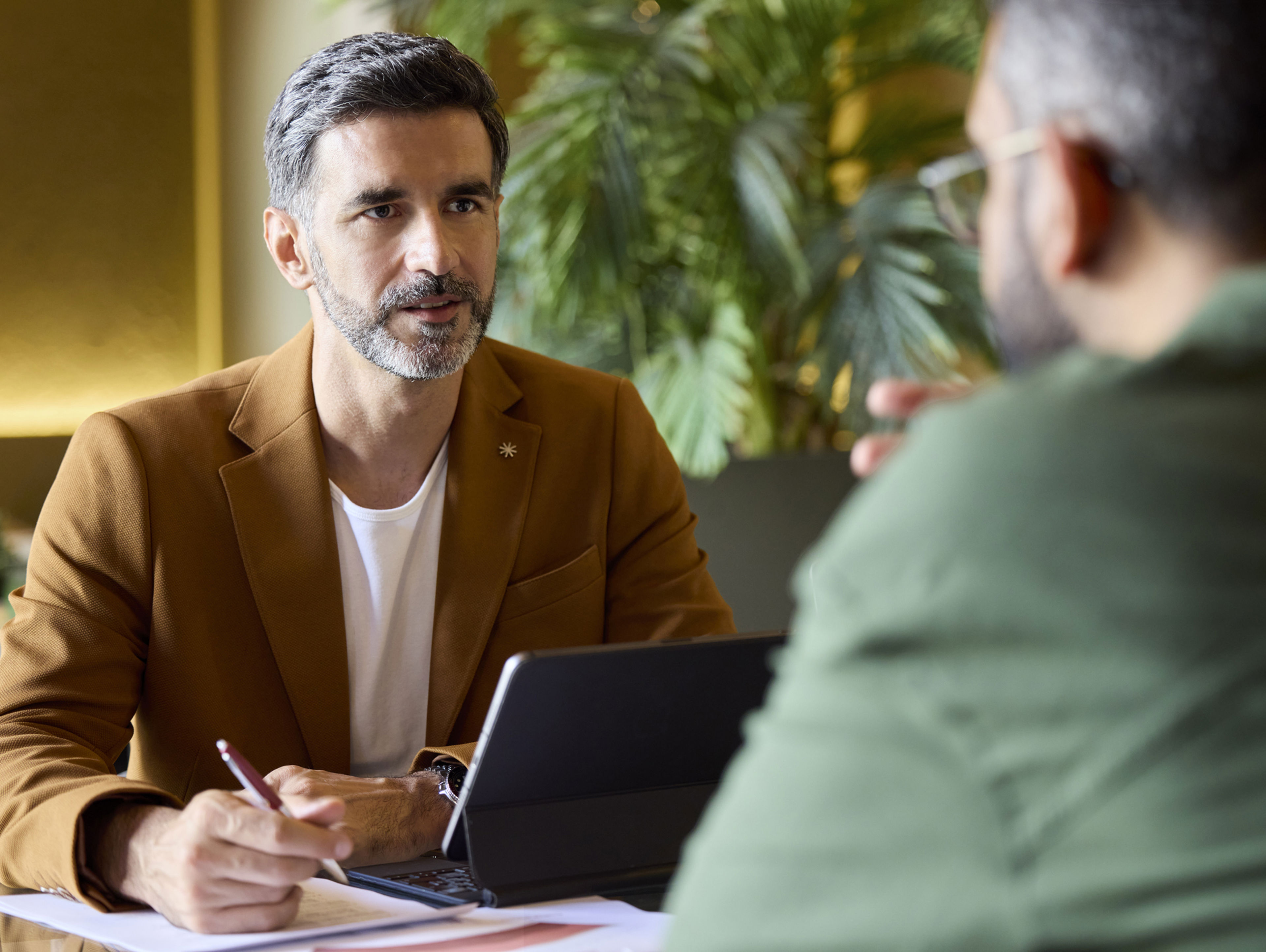 Two men have a serious discussion at a table with a laptop, notepad, and pen. One man in a brown jacket faces the camera, while the other, in green, sits with his back to the viewer. Indoor setting with plants visible.