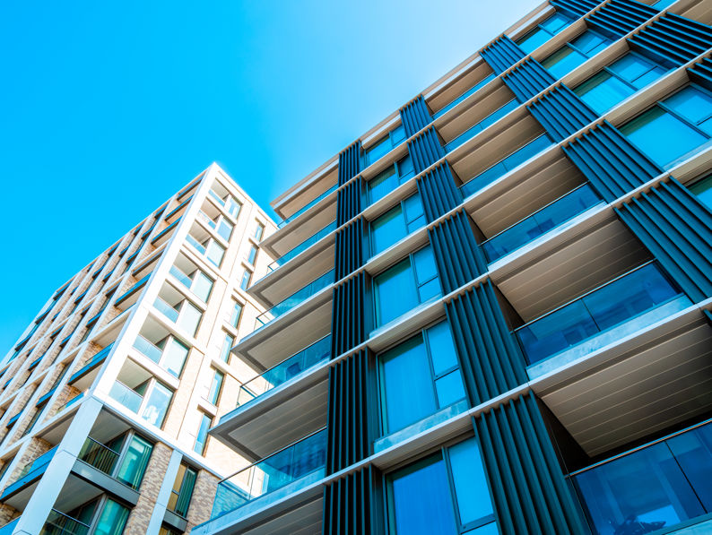 Modern apartment buildings with large glass windows and balconies, viewed from below against a clear blue sky. The architecture features clean lines and a mix of metal, glass, and brick elements.