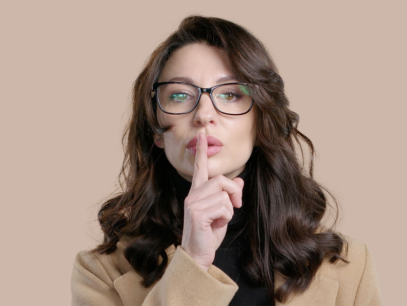 Woman with long brown hair and glasses wearing a beige coat, holding her index finger to her lips in a shushing gesture against a light brown background.