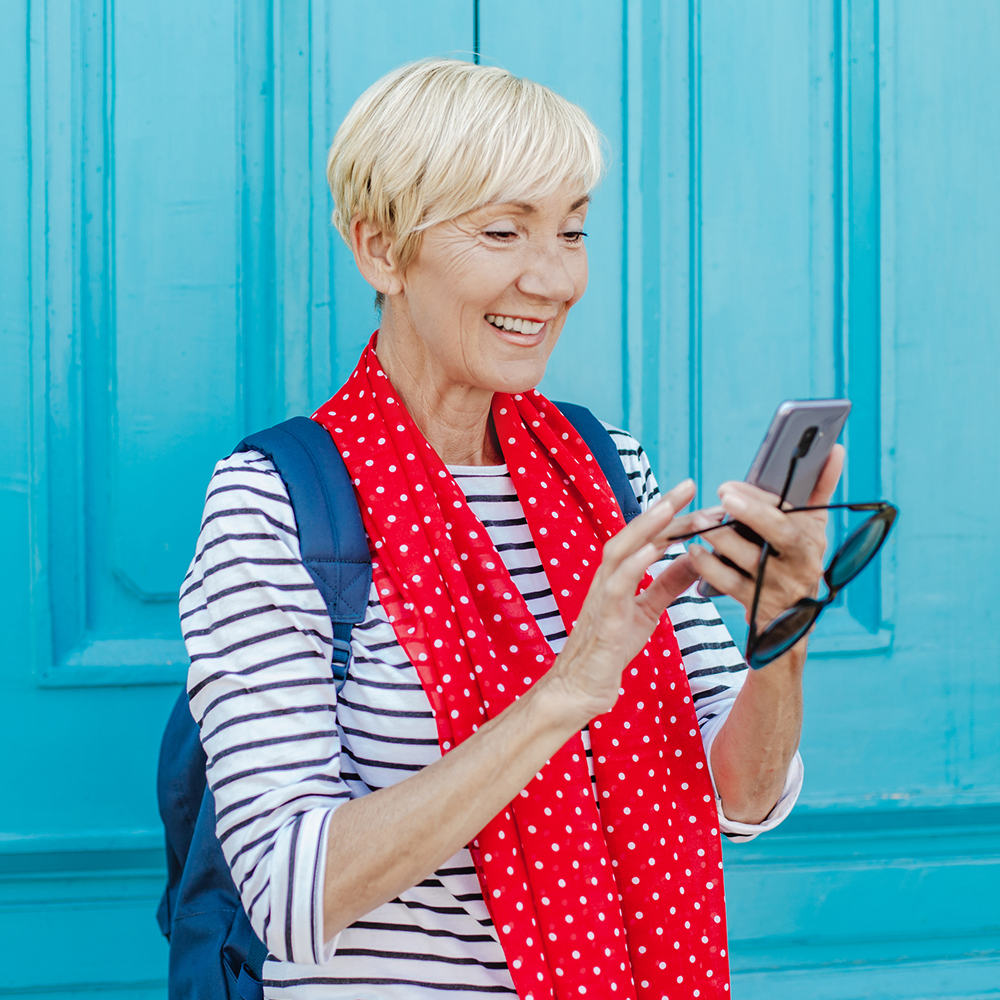 Smiling older woman with short blonde hair, wearing a striped shirt, red polka dot scarf, and backpack, holds a smartphone and glasses, standing in front of a bright blue door.
