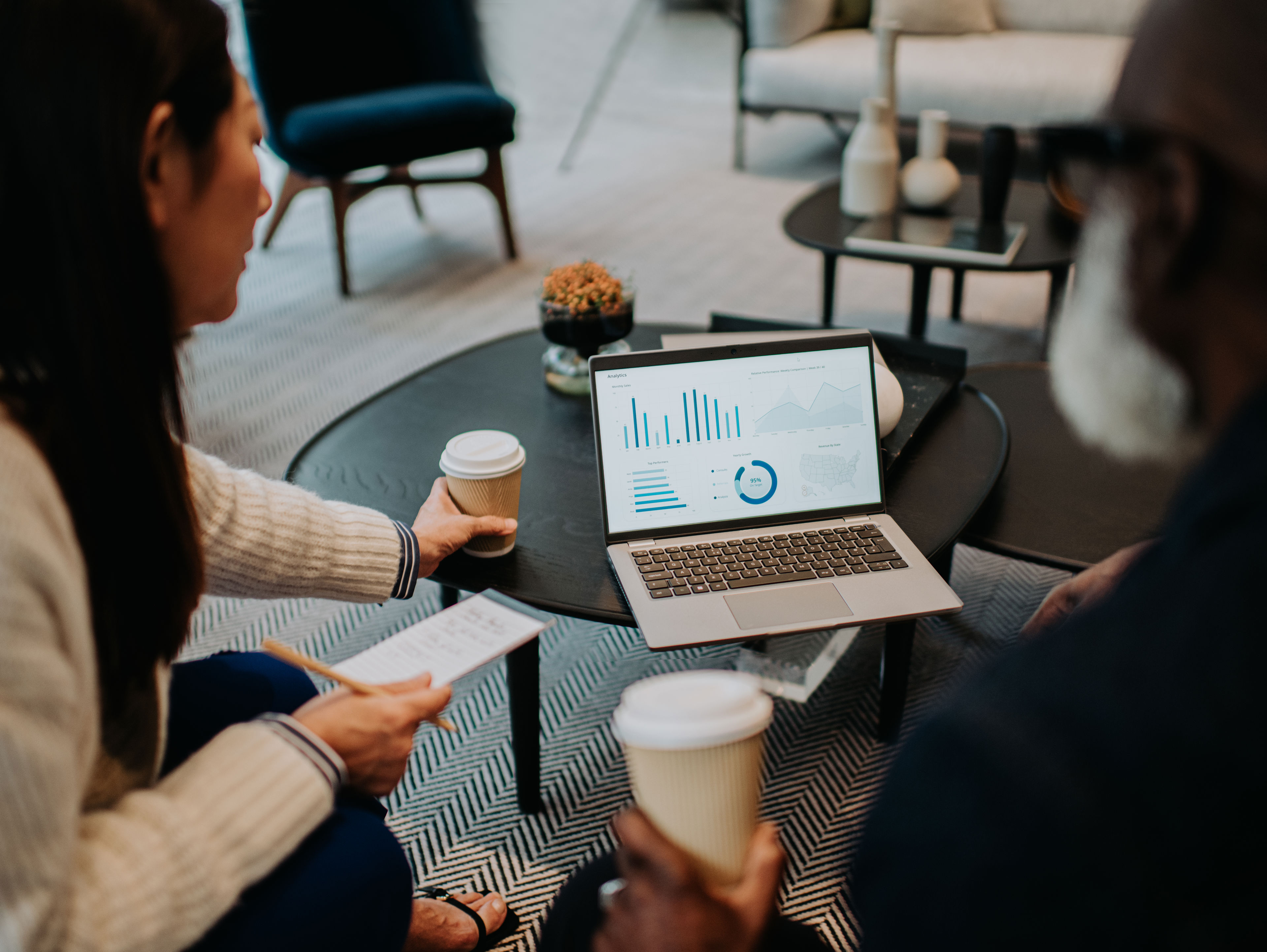 Two people sit at a coffee table with takeaway cups, discussing financial charts displayed on a laptop. One person holds a printed report, and the table has decorative items and a small potted plant.
