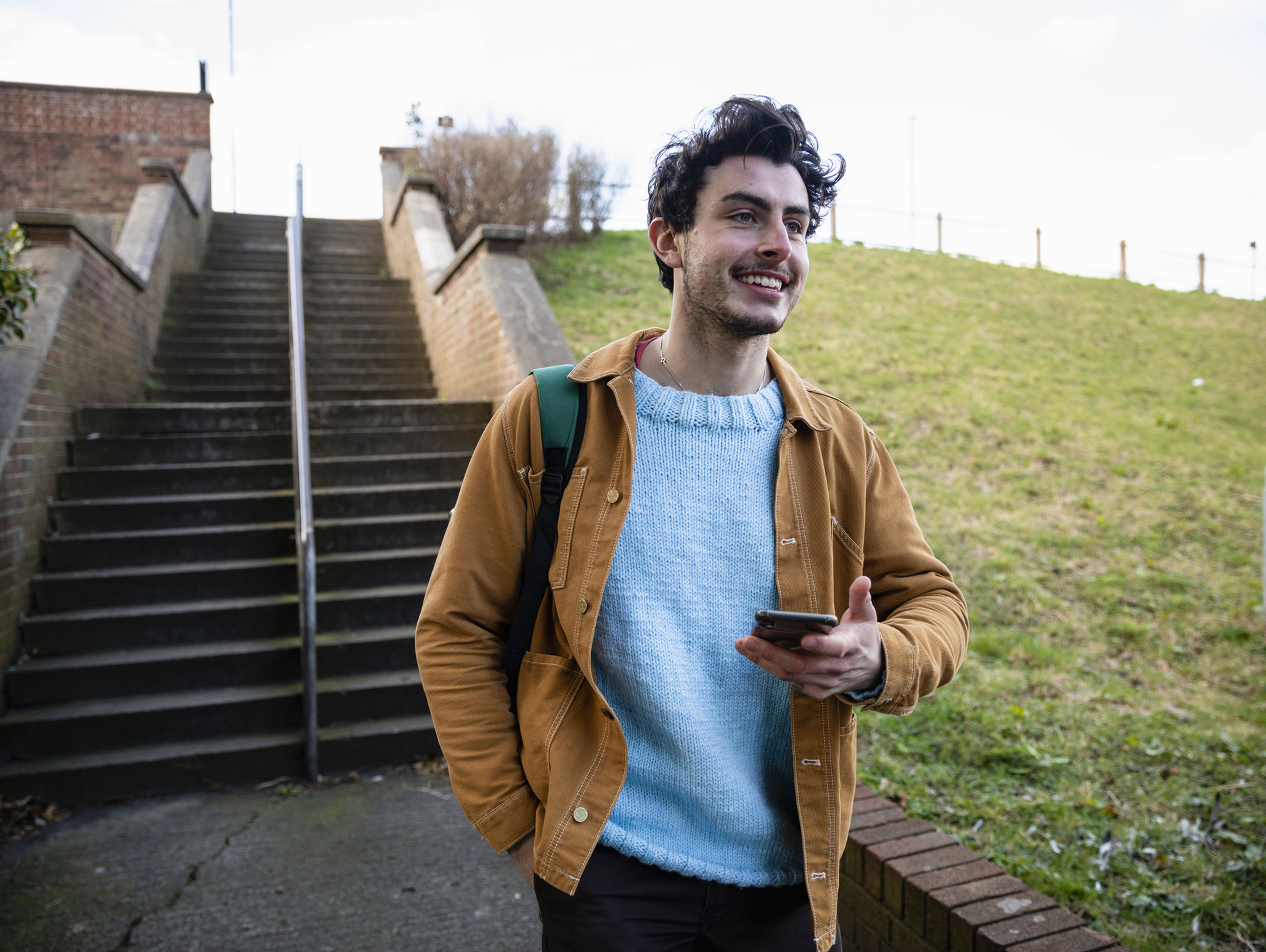 A person with dark hair smiles while standing outdoors near stairs. They are wearing a light blue sweater and a brown jacket, holding a smartphone, with a green backpack on one shoulder. Grass and a brick structure are in the background.