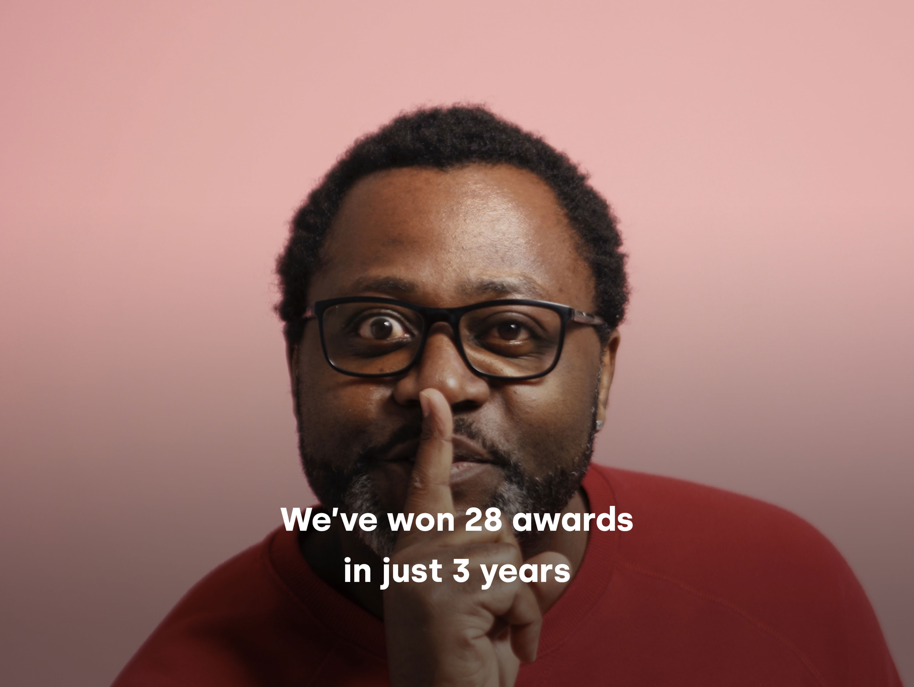 A man with glasses is holding a finger to his lips, signifying silence, against a pink background. He is wearing a red top. The text on the image reads, "We've won 28 awards in just 3 years.