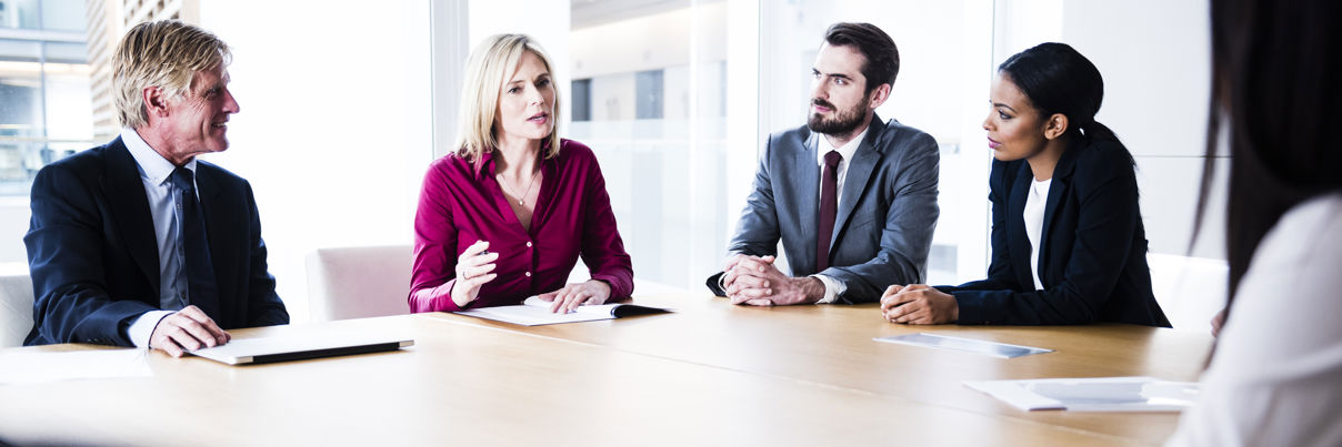 Five professionals sit around a conference table in a bright office, engaged in discussion. One woman in a red blouse is speaking while others, dressed in business attire, listen attentively.