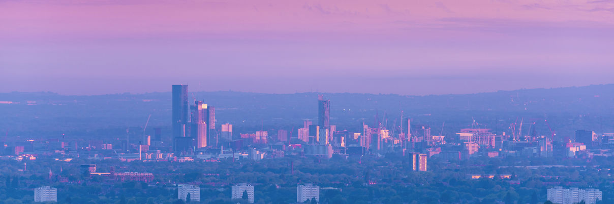 A cityscape at dusk with modern high-rise buildings and skyscrapers illuminated, set against a blue evening sky. Traditional historic buildings with domes and a clock tower are visible in the foreground.