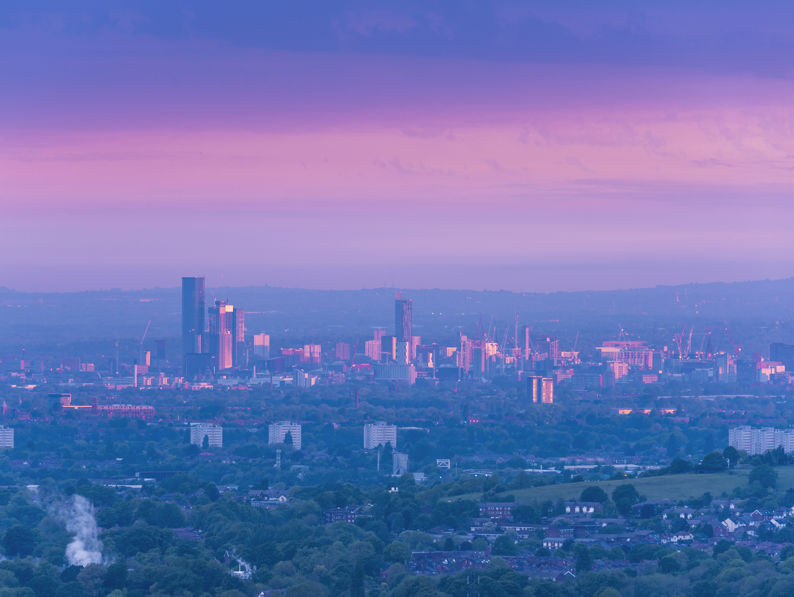 A cityscape at dusk with modern high-rise buildings and skyscrapers illuminated, set against a blue evening sky. Traditional historic buildings with domes and a clock tower are visible in the foreground.