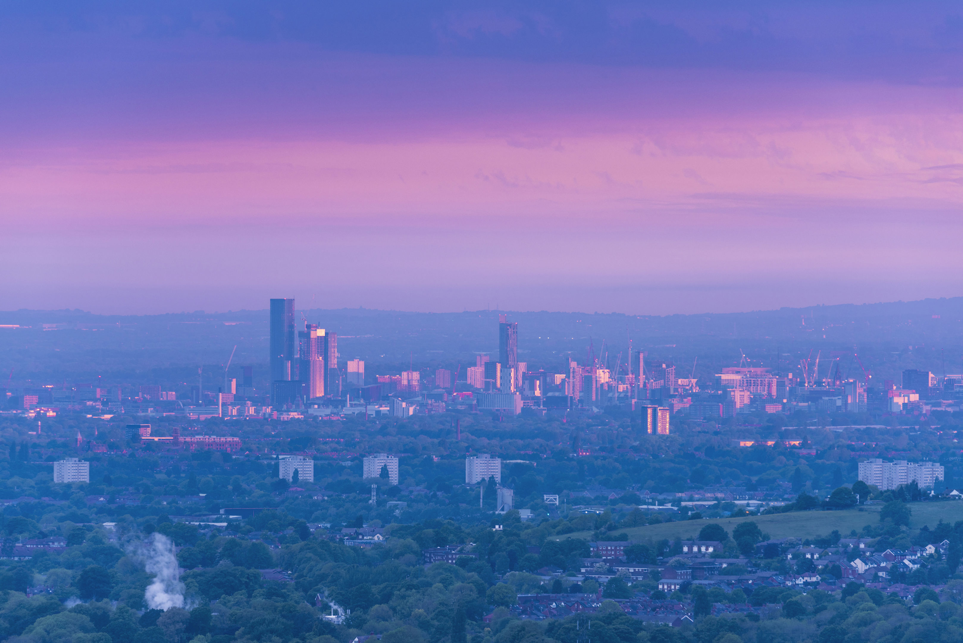 A cityscape at dusk with modern high-rise buildings and skyscrapers illuminated, set against a blue evening sky. Traditional historic buildings with domes and a clock tower are visible in the foreground.