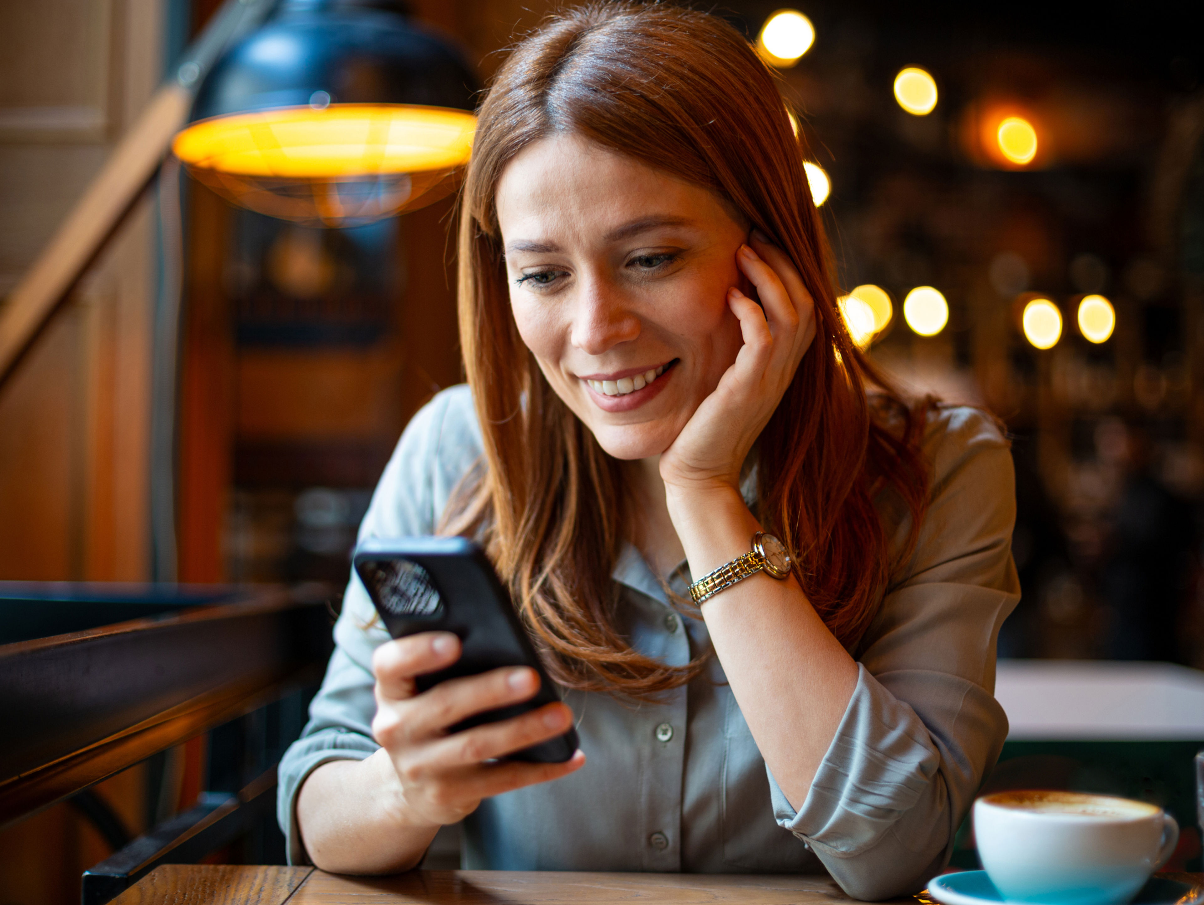 A woman with reddish-brown hair smiles while looking at her smartphone in a cozy cafe. She wears a light blouse and a watch, with a coffee cup on the table in front of her. Warm lights are blurred in the background.