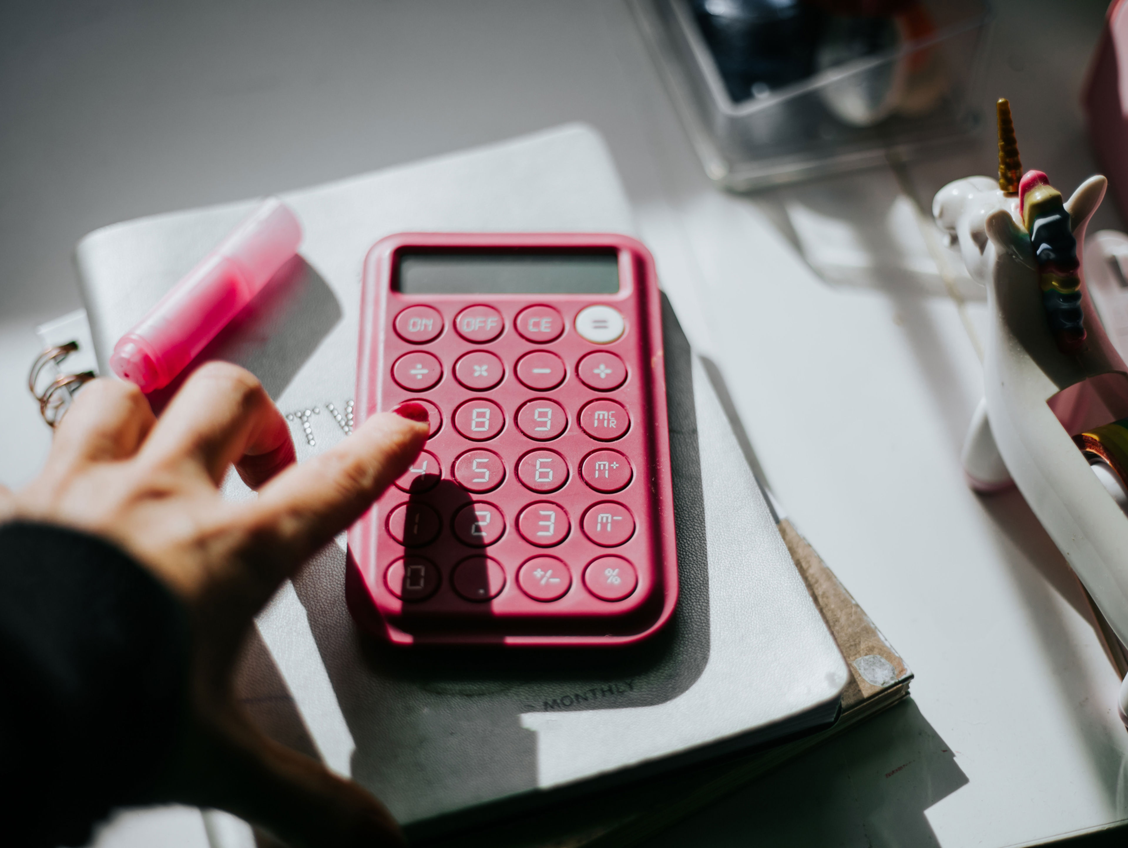 A hand pressing buttons on a pink calculator resting on a stack of notebooks. There is a pink highlighter on the left and a decorative tape dispenser shaped like a unicorn on the right. The scene is lit by natural light.