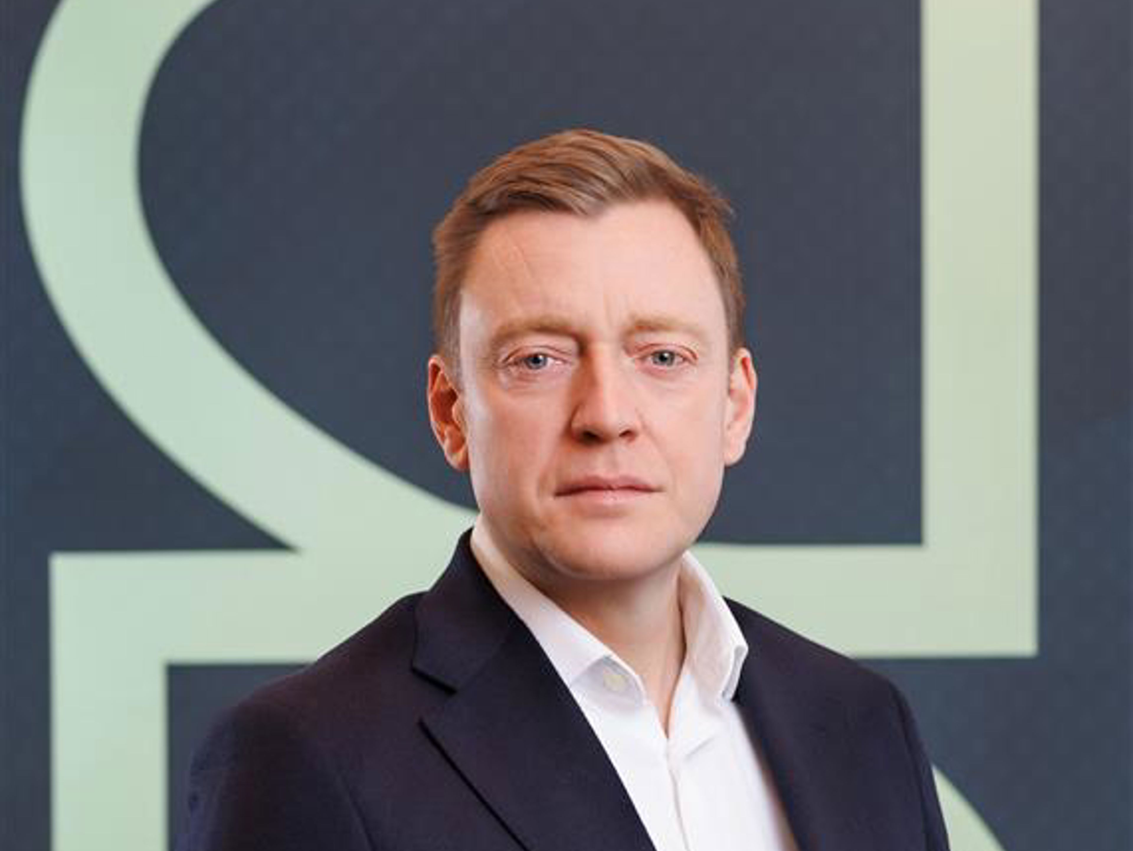 A man in a dark suit and white dress shirt stands in front of a geometric background featuring large, light-colored shapes. He is looking directly at the camera with a neutral expression.