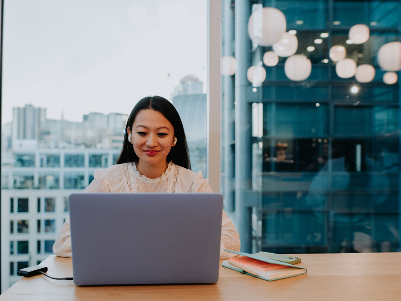 A young woman sits at a table in an office environment and uses a laptop - stock photo