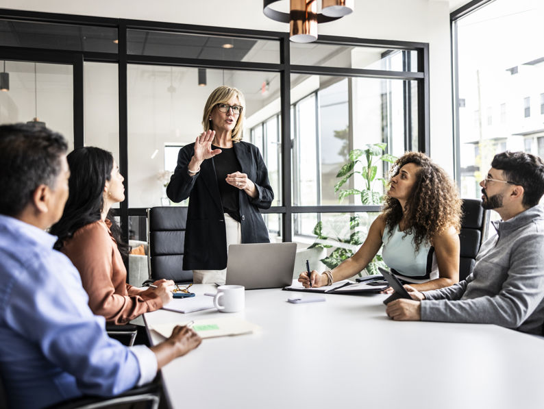 A woman stands addressing a group of four people seated around a conference table in a modern office. One person is taking notes, others listen attentively. A glass wall and potted plant are in the background.