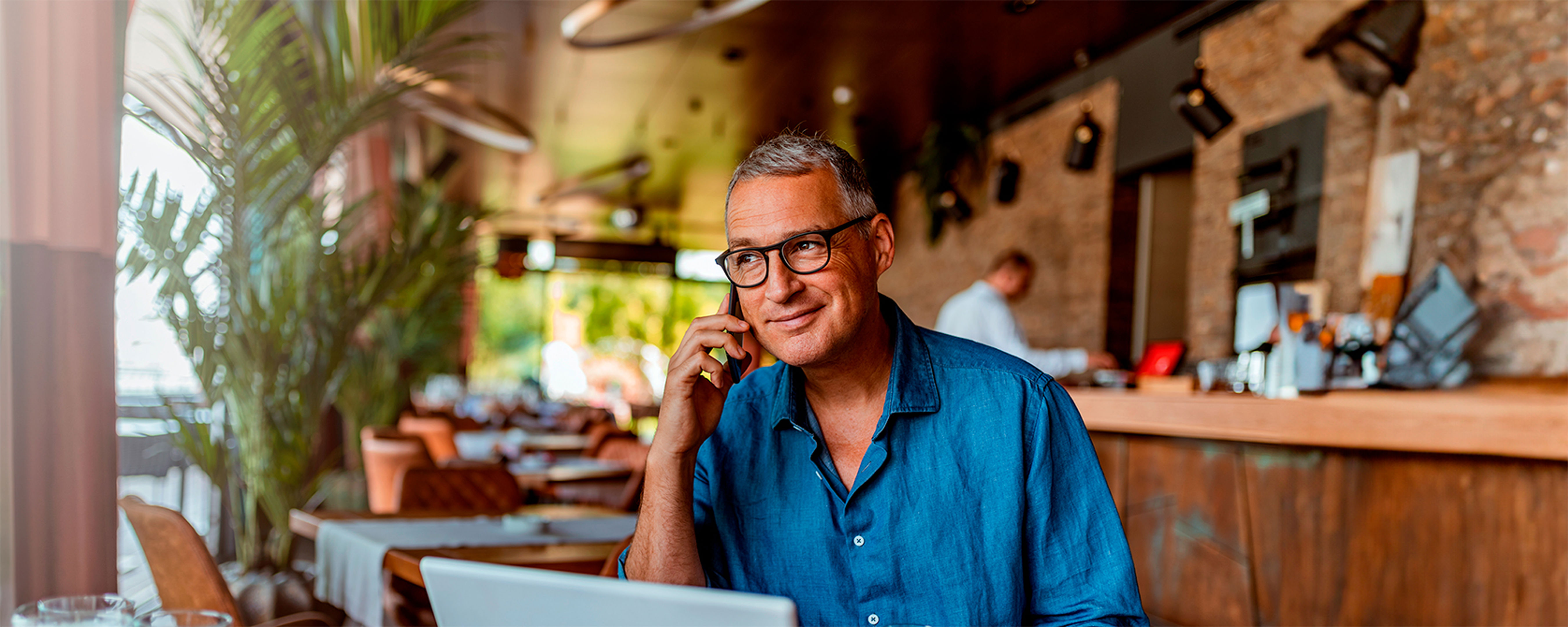 A man in a blue shirt is sitting at a cafe table with a laptop and a drink. He is smiling and talking on the phone. The cafe has wooden decor, shelves with bottles, and a row of tables. Lush green plants are in the background.