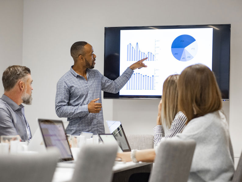 A man stands and points to graphs and charts on a screen during a business meeting, while four colleagues sit at a table watching and taking notes on laptops.