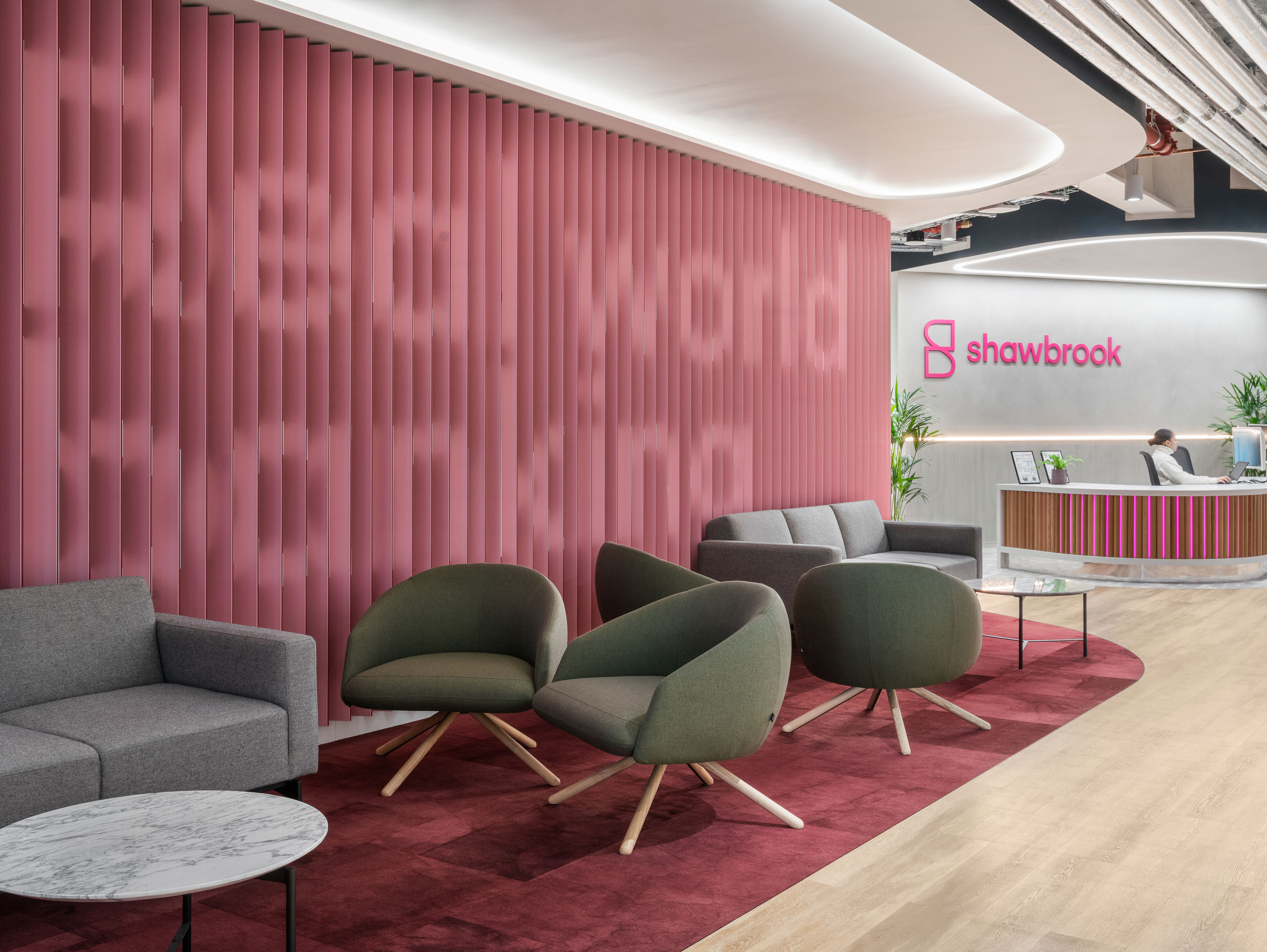 Modern office lobby with pink accent wall displaying "Real world banking," gray and green lounge chairs, a round marble table, and a reception desk with the Shawbrook logo in the background.