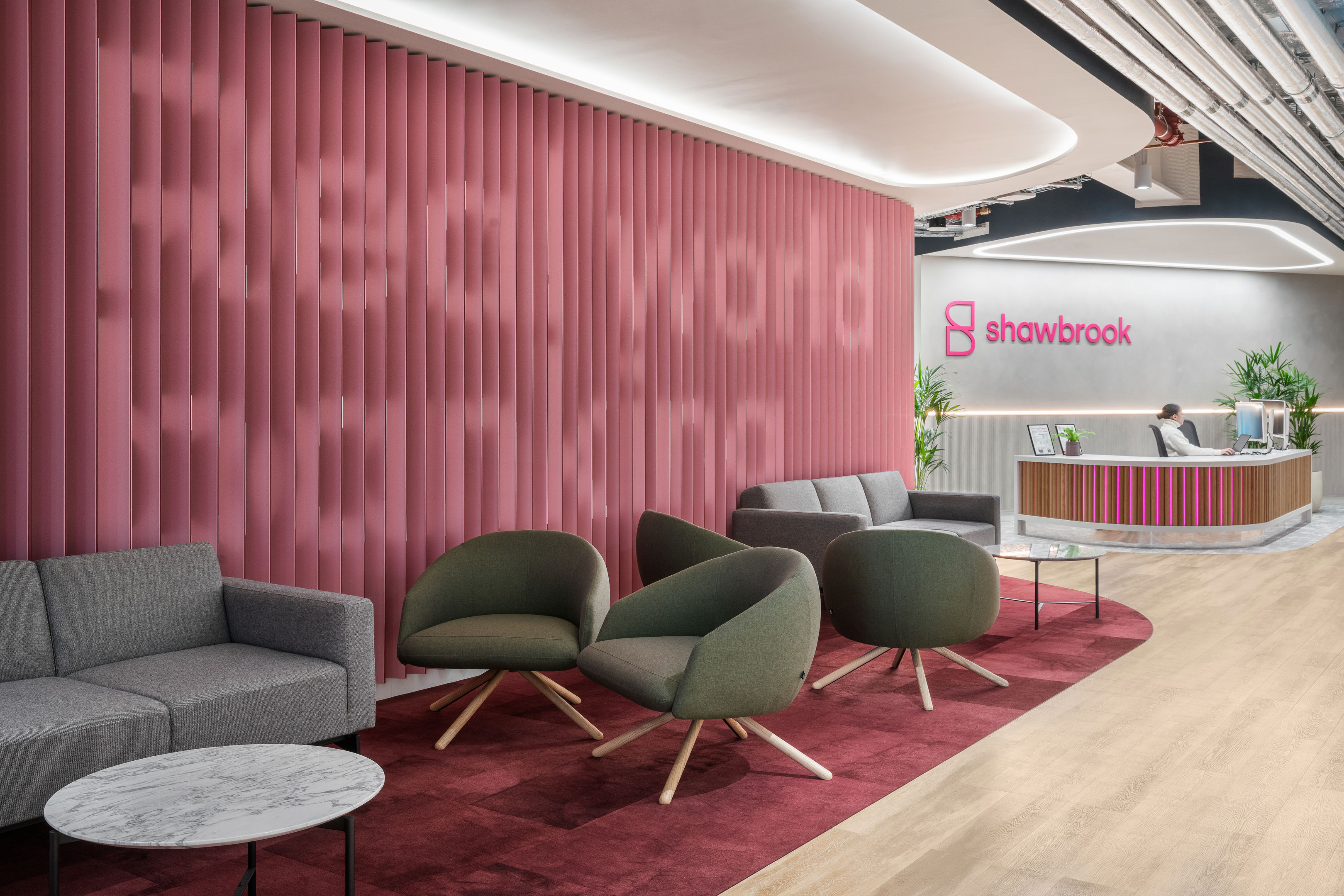 Modern office lobby with pink accent wall displaying "Real world banking," gray and green lounge chairs, a round marble table, and a reception desk with the Shawbrook logo in the background.
