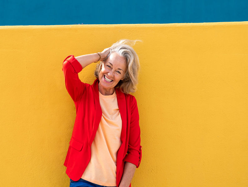 Smiling older woman with short blonde hair stands against a bright yellow wall, wearing a red blazer and peach shirt, with one hand behind her head. A teal section is visible above the wall.