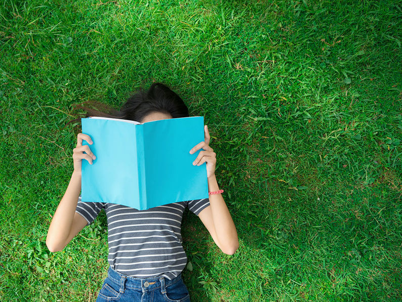 Woman Lying On Grass Reading Blue Book