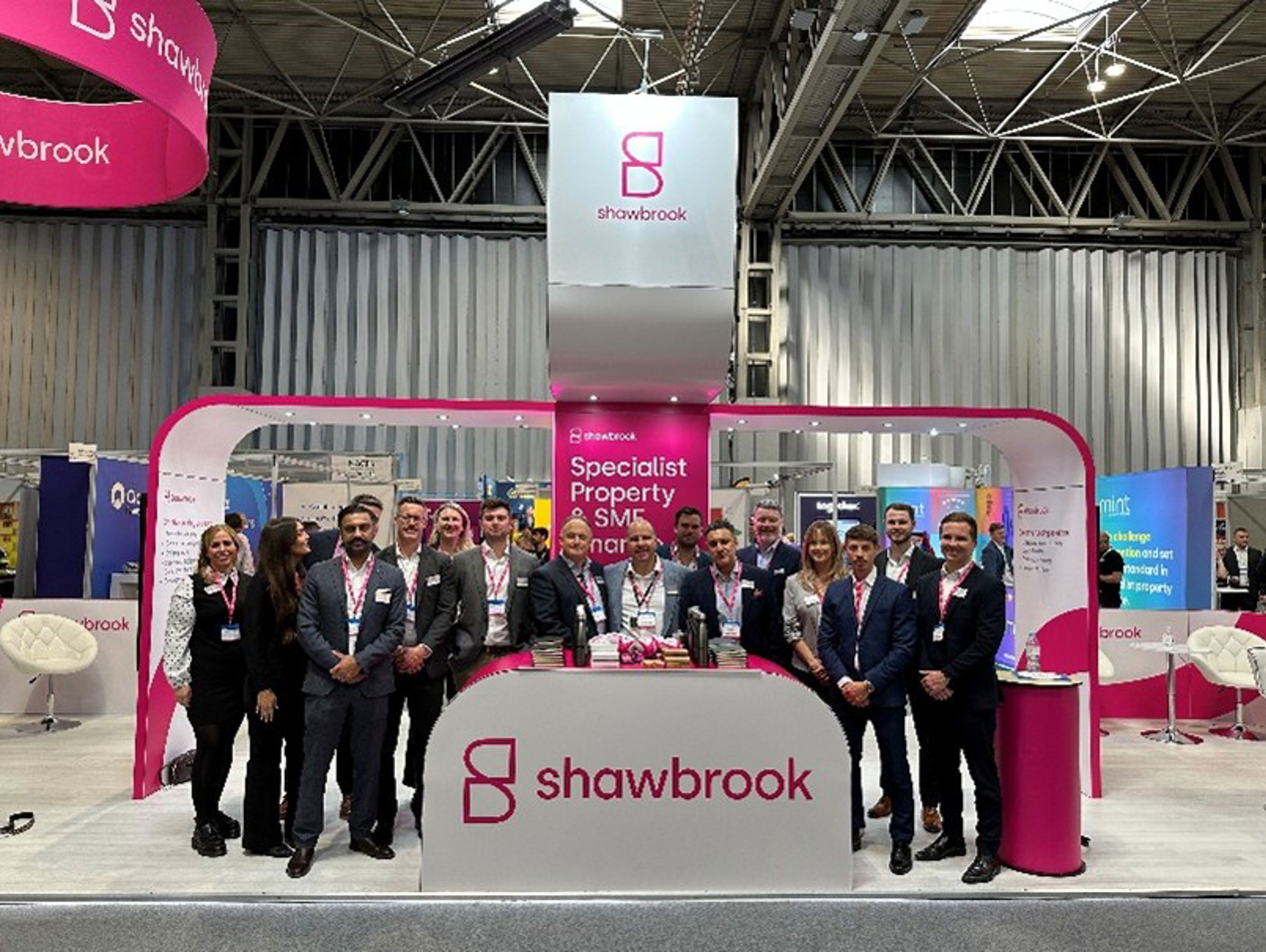 A group of professionals pose and smile in front of a Shawbrook exhibition booth, which features pink and white branding, a counter, and banners reading "Specialist Property" at an indoor event.