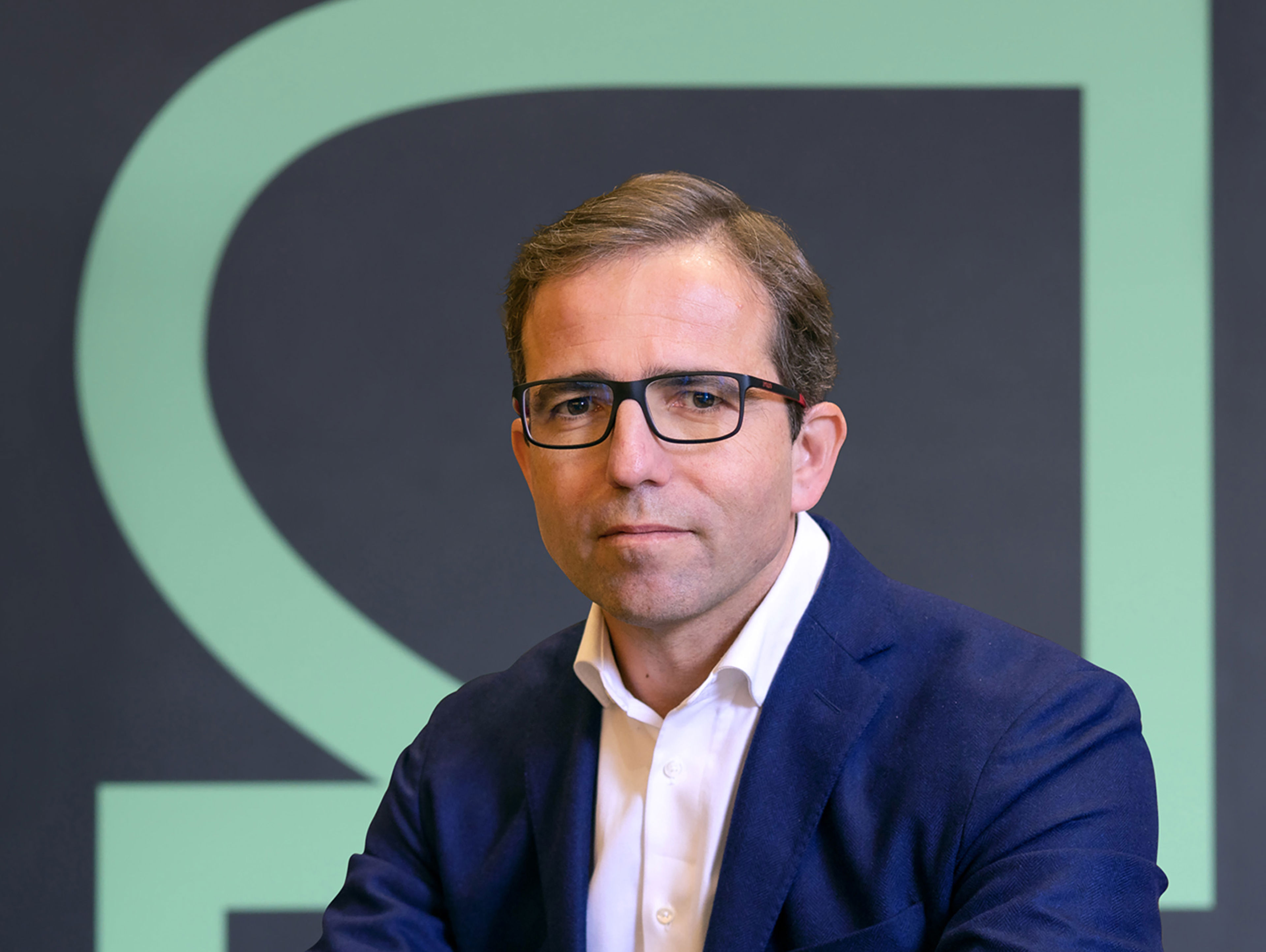 A man wearing glasses, a dark blue blazer, and a white shirt poses in front of a dark background with a large, light green geometric design.