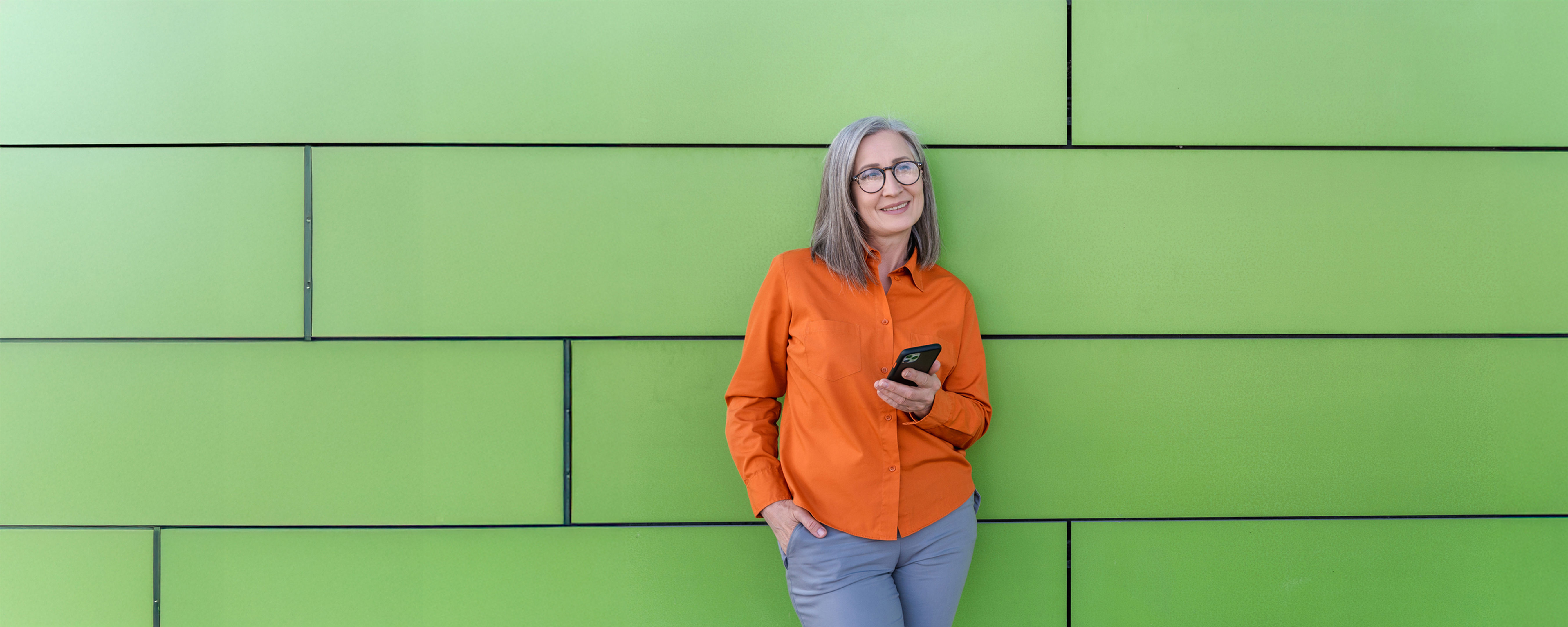 A gray tabby cat with green eyes lies on a white bed, looking up at the camera with its head resting on a pillow and one front paw curled near its face.